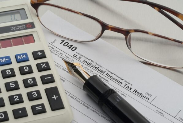 A close-up image of a 1040 U.S. Individual Income Tax Return form. A pair of eyeglasses, a fountain pen, and a calculator are placed on top of the form, suggesting tax preparation and calculation activities.
