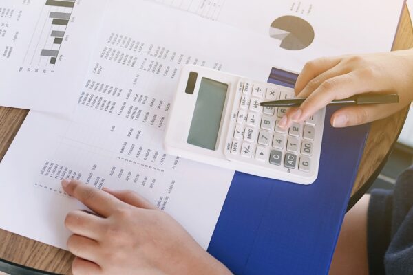 A person is using a white calculator on a table, surrounded by printed sheets with charts, graphs, and numeric data. One hand is pressing a button, and the other hand is pointing at a section of the paper. The calculator sits on a blue folder.