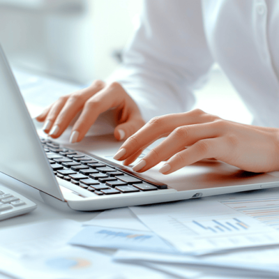 A person types on a laptop keyboard, surrounded by sheets of paper with graphs and data. The scene conveys a work or study environment, focused on productivity and analysis.